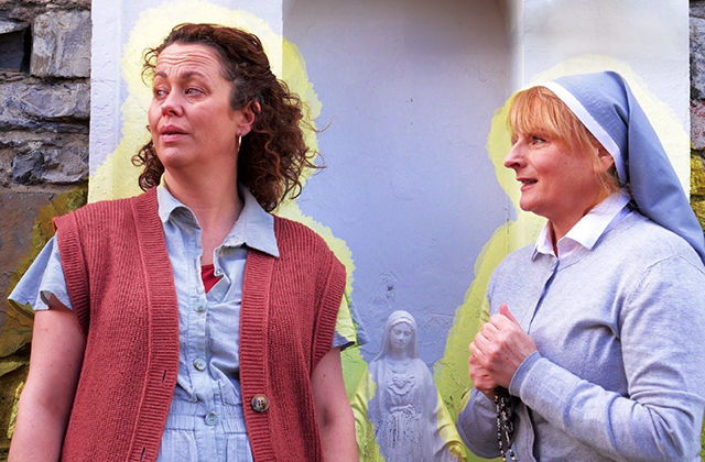 Poster for Philo, Neilí Conroy in a red cardigan and Niamh McGrath, dressed as a nun, are both facing right with a bright yellow glow around them, and a statue of the virgin Mary in the background.