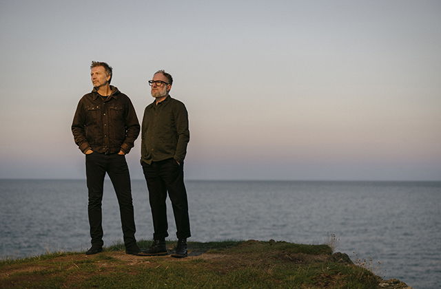 Paul Noonan and Brian Crosby stand on a cliff's edge with the sea behind them