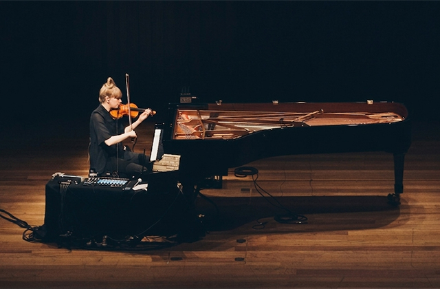 Solo violinist performs beside a grand piano on a dimly lit stage, illuminated by a warm spotlight against a dark auditorium backdrop.
