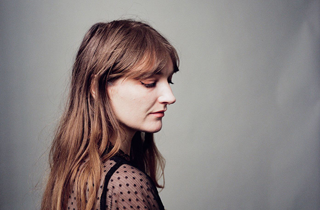 Portrait of a Aoife Wolf in profile, a woman with long light-brown hair, wearing a sheer black polka-dot top against a soft grey background.