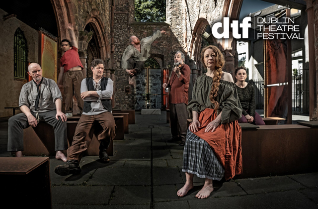 Seven performers are posed inside the roofless stone ruins of a church or abbey, surrounded by Gothic arches and weathered walls. A barefoot woman with long red hair sits prominently in the foreground wearing layered period-style clothing in rust, grey and olive tones. Other cast members are seated, standing or leaning throughout the space, creating a dramatic tableau. The scene is lit with theatrical contrast, highlighting the historic architecture. The Dublin Theatre Festival logo appears in the corner.