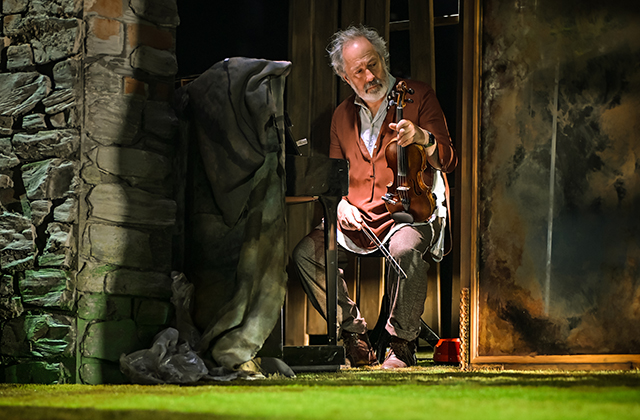 A performer sits on a chair at the edge of a grassy stage, holding a violin and bow. The performer wears layered, rustic clothing including a brown cardigan. Behind, a stone wall and vertical wooden panels frame the set, with a draped fabric and a painted backdrop to the side. Warm theatrical lighting highlights the scene, with a small red object on the ground nearby.