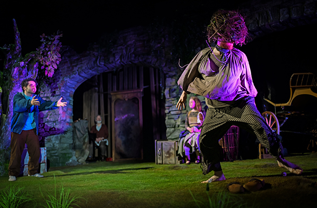 A performer sits on a chair at the edge of a grassy stage, holding a violin and bow. The performer wears layered, rustic clothing including a brown cardigan. Behind, a stone wall and vertical wooden panels frame the set, with a draped fabric and a painted backdrop to the side. Warm theatrical lighting highlights the scene, with a small red object on the ground nearby.