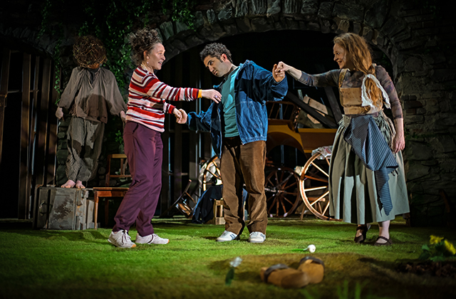 Three performers stand on a grassy stage holding hands in a loose circle. One in a blue jacket stands between two others, arms outstretched. Behind them, two more performers stand near stone archways covered in greenery, with a wooden cart and scattered furniture. Soft theatrical lighting illuminates the scene, with small props visible on the ground.