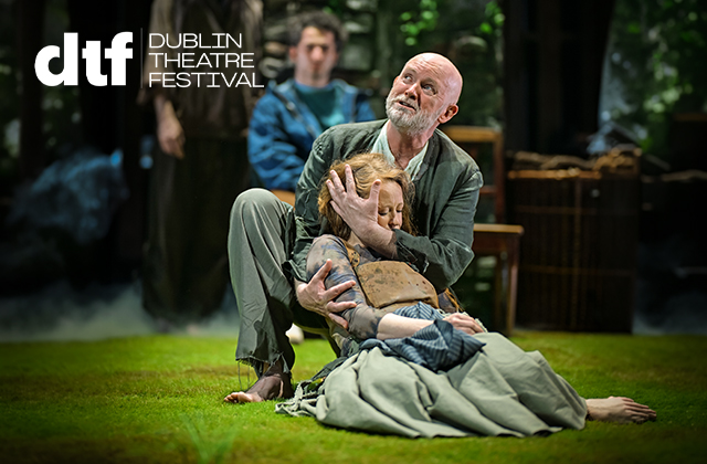 Performer kneels on a grassy stage, supporting another performer lying wrapped in layered, earth-toned clothing and a blanket. The kneeling performer cradles the other’s head. A third performer and wooden set pieces appear blurred in the background with soft, misty lighting. White text in the top left reads “dtf” and “Dublin Theatre Festival.”