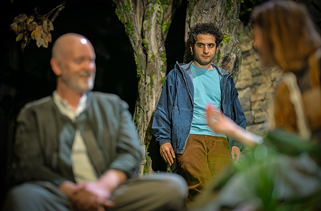 A performer in a blue jacket and brown trousers stands beside a tree trunk on a grassy stage, facing another performer in the foreground who is out of focus. A third performer sits blurred in the left foreground. The set features stone archways covered in greenery and soft theatrical lighting, with plants and branches framing the scene.