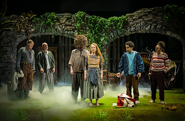 Six performers stand on a grassy stage with low mist, framed by stone archways covered in greenery. At centre, one performer faces forward wearing layered, earth-toned clothing, while another beside them has a large, textured headpiece obscuring the face. Additional performers stand to either side in casual, muted outfits. Scattered props include a tipped chair, a bag on the ground, and wooden items, all lit by soft, theatrical lighting.