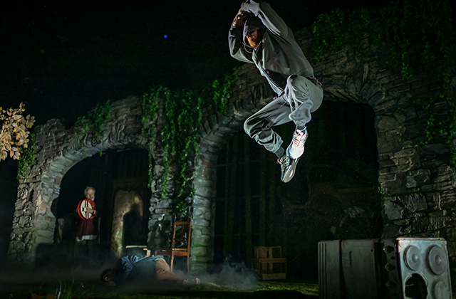 Performer kneels on a grassy stage, supporting another performer lying wrapped in layered, earth-toned clothing and a blanket. The kneeling performer cradles the other’s head. A third performer and wooden set pieces appear blurred in the background with soft, misty lighting. White text in the top left reads “dtf” and “Dublin Theatre Festival.”