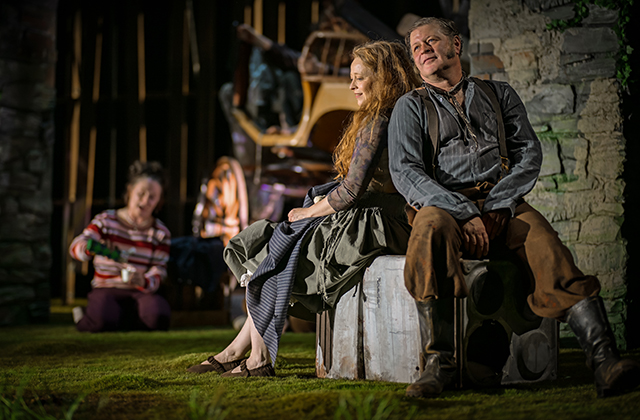 Two performers sit back-to-back on a low platform on a grassy stage, wearing layered, earthy-toned costumes. Behind them, a third performer kneels on the ground holding a small object. The background includes stone walls, vertical wooden elements, and stacked furniture pieces such as chairs. Warm theatrical lighting highlights the scene against a darker backdrop.