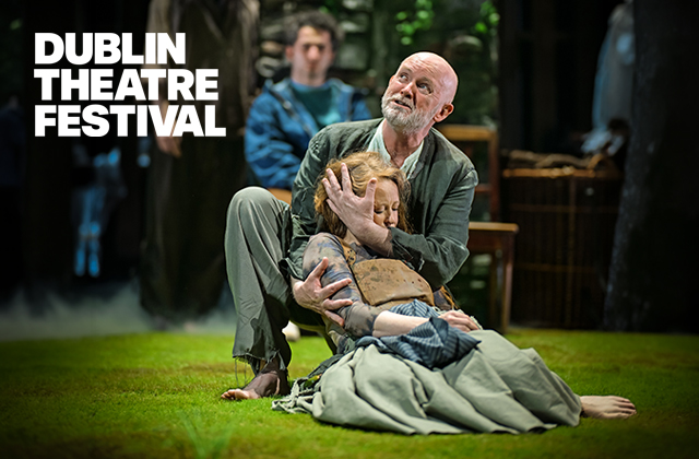 Performer kneels on a grassy stage, supporting another performer lying wrapped in layered, earth-toned clothing and a blanket. The kneeling performer cradles the other’s head. A third performer and wooden set pieces appear blurred in the background with soft, misty lighting. White text in the top left reads “dtf” and “Dublin Theatre Festival.”
