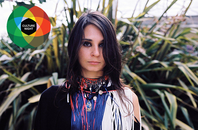 A woman with long dark hair looks directly forward, wearing an intricate statement necklace with small metallic coins and a vibrant, fringed garment with red, blue, and white tassels. Lush, spiky tropical foliage fills the outdoor background. In the upper left corner, a circular logo made of overlapping colored circles surrounds a black center with the text