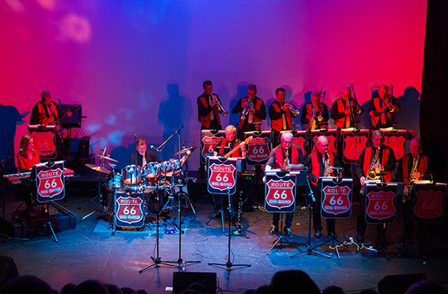 A live performance by the Route 66 Big Band on stage. The band members, dressed in coordinated black and red outfits, play various instruments including drums, keyboards, saxophones, trumpets, and trombones. Each music stand displays the Route 66 logo. The stage is lit with vibrant red and blue lighting, creating a dynamic and colourful atmosphere.
