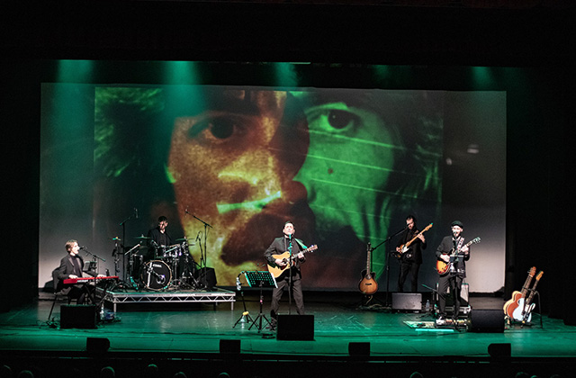 A wide shot a band on stage fronted by Daniel Taylor as George Harrison with an image of George as a backdrop projected behind.