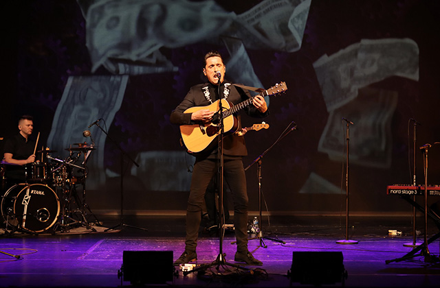 A close up of Daniel Taylor performing as George Harrison in There's Something About George. Daniel stands on stage in a black suit, holding his guitar and singing.