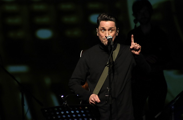 Another close up of Daniel Taylor as George Harrison. Taylor stands to the right of the image, he is in a black suit with the guitar strapped around his side, he is looking intensely towards the camera with his finger pointing upward.