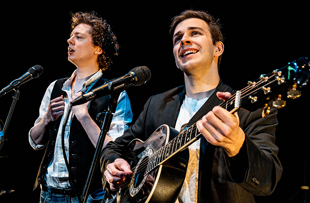 Close-up of two young male musicians performing on stage. The performer on the right plays an acoustic guitar and smiles, while the singer on the left performs into a microphone.