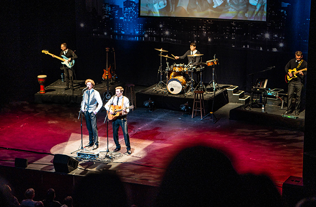 Wide view of a live band performing on stage in front of a backdrop showing a city skyline illuminated by stage lights. Two singers stand front center while a bassist, drummer, and guitarist perform behind them.
