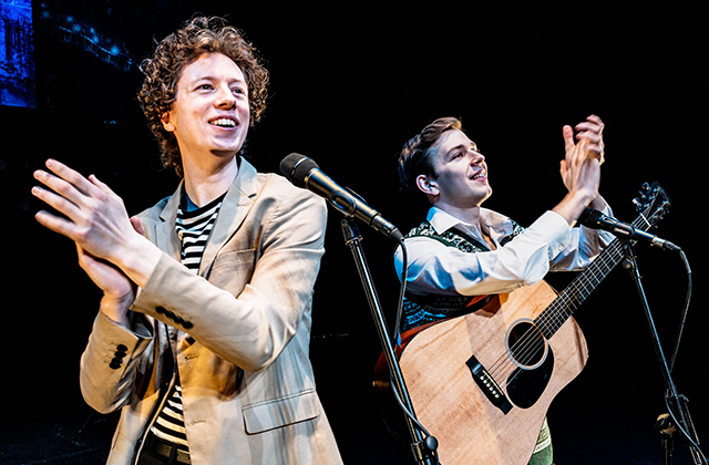 Close-up of two male performers on stage smiling and clapping hands towards the audience, with microphones and an acoustic guitar visible in the foreground.