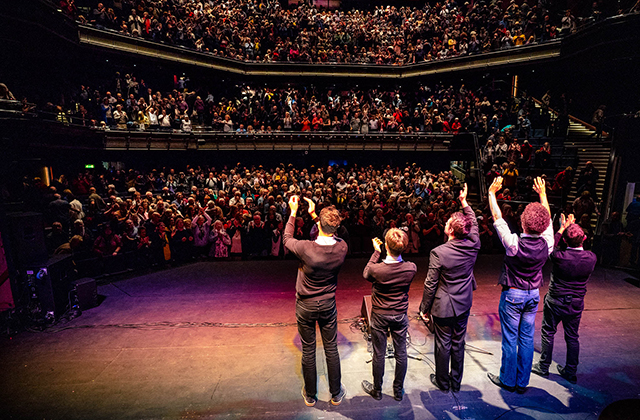 View from behind five band members standing at the edge of a stage with hands raised, waving to a large, packed multi-tiered theater audience applauding at the end of a performance.