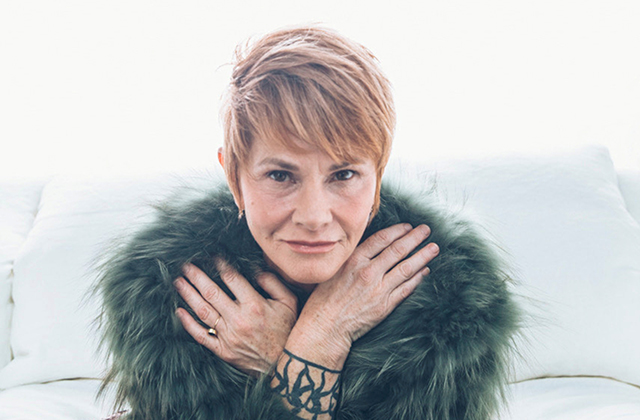 Shawn Colvin, a woman with short blonde hair, resting her crossed hands near her face, wearing a dark green fur wrap and showing a tattoo on her wrist against a pale background.