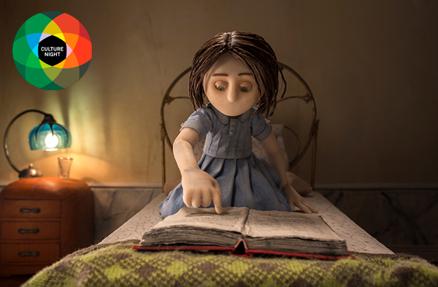 Image from 'Ana Morphose' - A stop-motion young girl with black hair, and a blue dress, is sat on a small bed pointing at a book