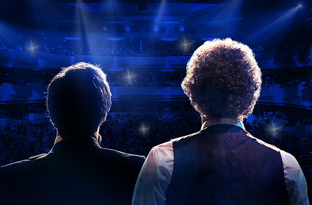 Two performers dressed as Simon and Garfunkel with their backs to the camera, staring out to a packed theatre