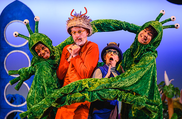 2 performers, one in blue and one in red, are surrounded by long green arms in a big hug