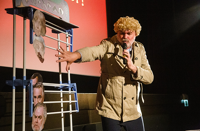 Tim Benzie in a blonde wig and tan trenchcoat, gesticulating at a board with cut-outs of actors' faces stuck to it.