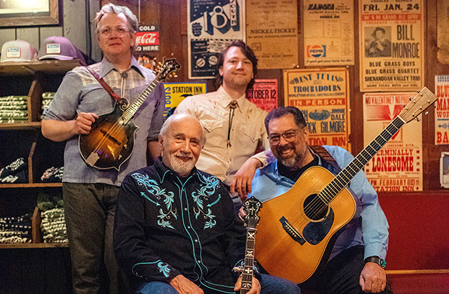 Members of Special Consensus gather with their instruments for a group portrait in a rustic bluegrass venue surrounded by vintage performance posters.