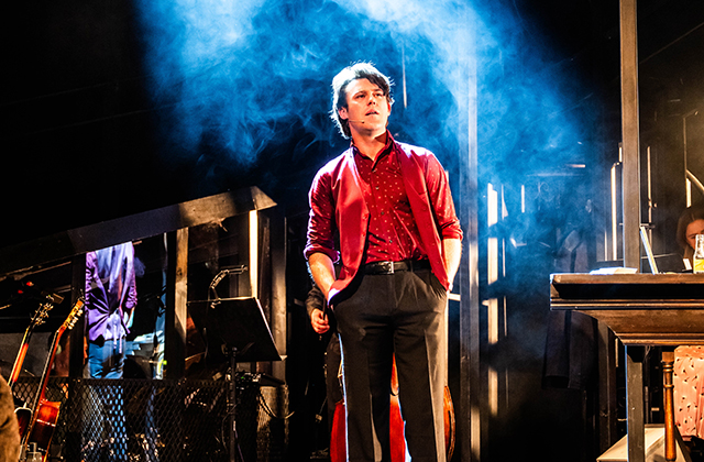 Niall McNamee from The House Must Win. He stands centre stage in a read waistcoat and t-shirt. he looks out towards the audience