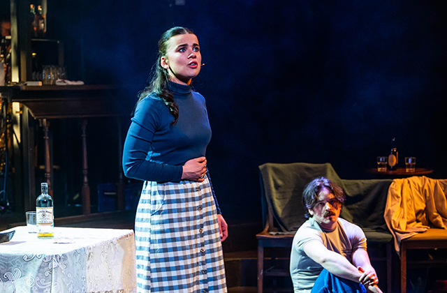 Tabitha Smyth stands in a blue polo neck and blue and white patterned dress. She looks worried and stares out away to the distance.