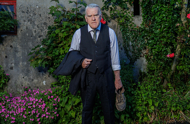 Mikel Murfi stands hunched in the garden of a rural cottage, dressed in a suit, and holding a battered shoe