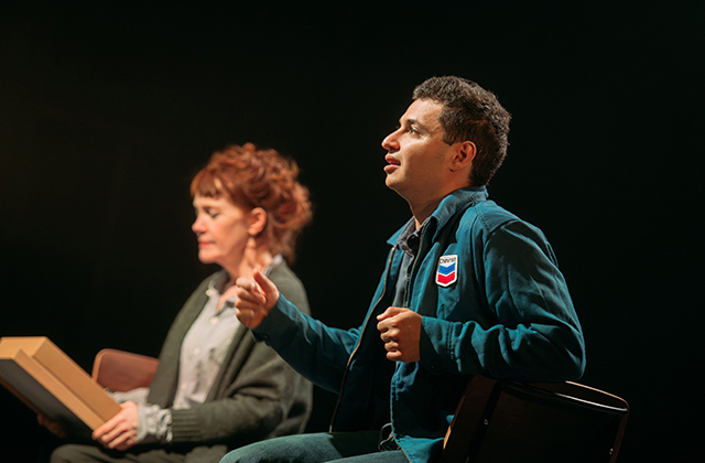Still from production - An older woman in the background, while a younger man sits facing forwards speaking animatedly.