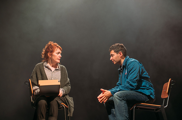 Still from production - An older woman sits facing out, with books on her lap, while a younger man sits facing sideways gesturing towards her