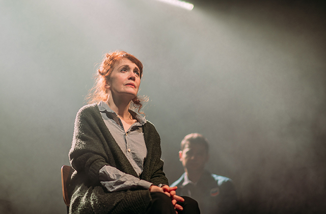 Still from production - An older woman sits facing out, with the figure of a younger man standing behind her