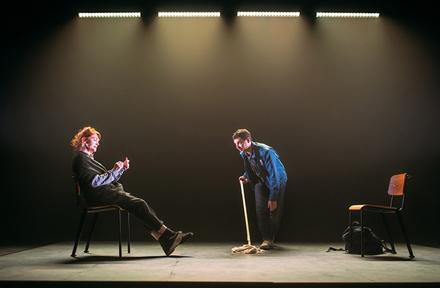 Still from production - a woman sits stage left, while a younger man stands centre stage facing her holding a mop