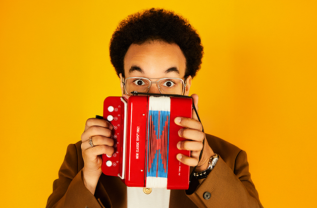 A close up shot of Théo Ould, he is pictured against a bright yellow background, holding up a small bright red and blue mini accordion, which covers the lower part of his face. He is wearing a glasses and he his eyes are open wide giving a surprised expression.