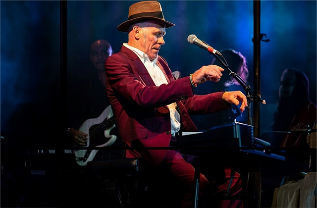 Stewart D’Arrietta sits at a piano wearing a red suit, and brown hat