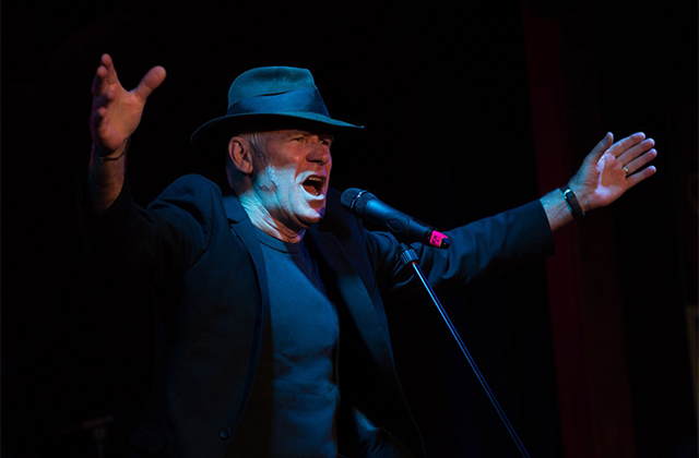 Stewart D’Arrietta dressed in black, with a black hat, sings into a microphone with his hands outstretched