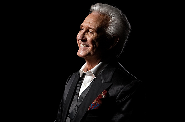 A portrait of Tony Christie, smiling looking off camera, wearing a dinner suit, with the bow tie undone