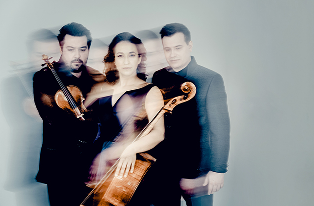 Trio Bohémo are picture together, from left Matouš Pěruška holding fiddle, Kristina Vocetková holding a cello, Jan Vojtek stands they are all wearing black evening attire and the image is blurred in an artistic style