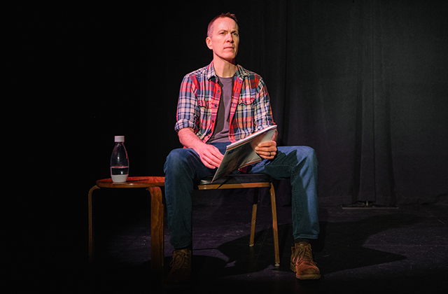 A man wearing a red-and-blue plaid shirt and jeans sits on a chair on a dim stage, holding a booklet or manuscript. A small side table with a clear water bottle stands beside him against a black backdrop.