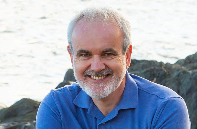 A close-up portrait of a Colm O'Gorman, a smiling middle-aged man with short grey hair and a full grey beard, wearing a blue polo shirt with a body of water and rocky coastline in the background.