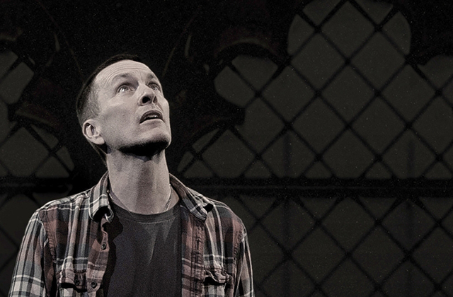 A man in a plaid shirt and jeans stands atop a wooden chair on a dark stage with his arms outstretched. Beside him, a framed projection screen displays a religious scene with a crucifix and figures in a church.