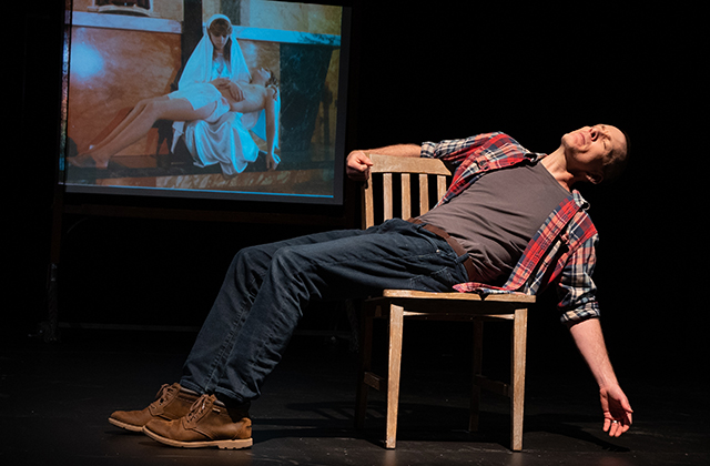 Jay Sefton lays back sideways on a wooden chair, with his left arm outstretched. An image of the Pieta is projected behind him, and you can see that Jay is holding the same pose as Jesus