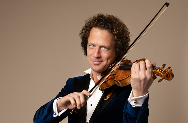 Vladimir, a violinist with curly hair wearing a dark velvet tuxedo jacket, plays a violin, holding the bow mid-stroke and looking toward the camera against a plain beige background.