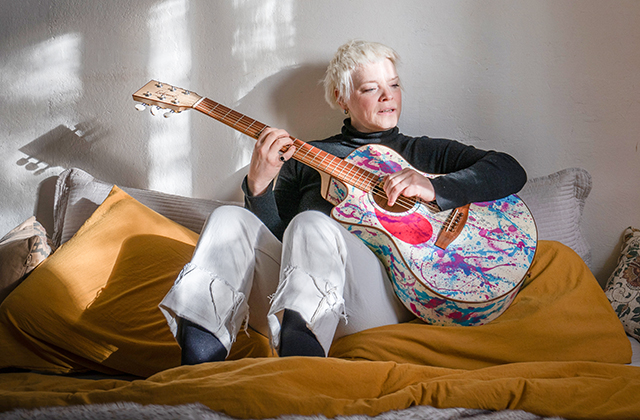 Wallis Bird, a woman with short light hair, sitting on a bed or sofa, playing an acoustic guitar decorated with colourful paint splashes. Sunlight streams across the wall and cushions behind her, creating soft shadows, while she looks down at the guitar as she plays.