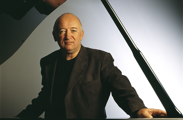 John O'Connor stands in front of his grand piano looking towards the camera with a pensive expression on his face. He is wearing a black shirt and jacket.