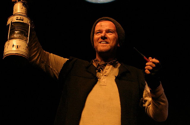 The photo depicts Aidan Dooley as Tom Crean. A man stands towards the camera, the moon is above him, one arm is raised holding up a lamp, the other arm is half raised holding a pipe. He is dressed in a hat and tunic.